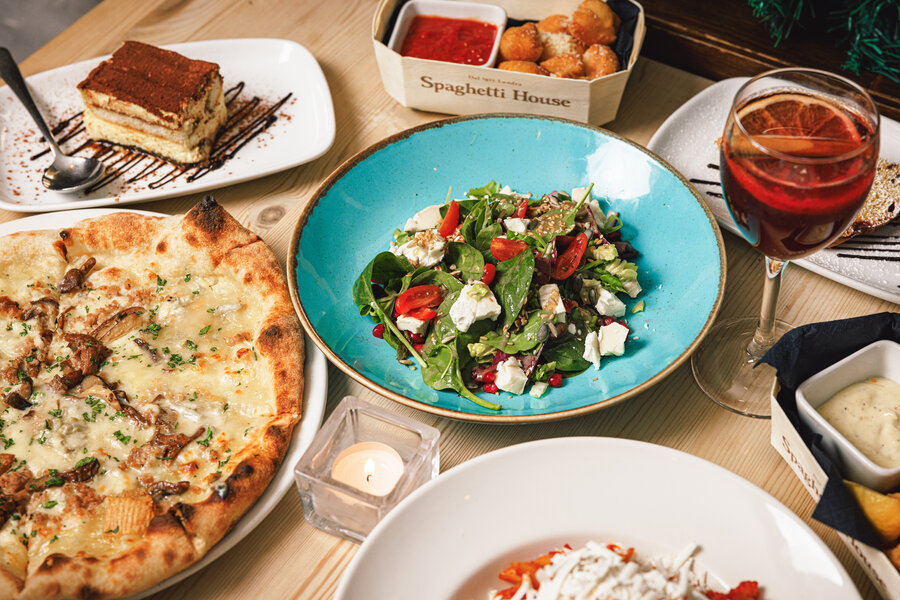 A table with a variety of dishes: a cheesy pizza, a fresh salad with feta and cherry tomatoes, a dessert of tiramisu, fried snacks with dipping sauce, and a glass of red wine.