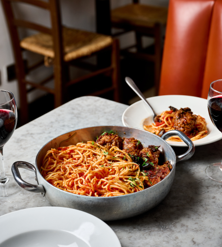 A metal pot filled with spaghetti and meatballs garnished with herbs sits on a marble table. In the background, a white plate with a smaller portion and two glasses of red wine are visible, along with a wooden chair.